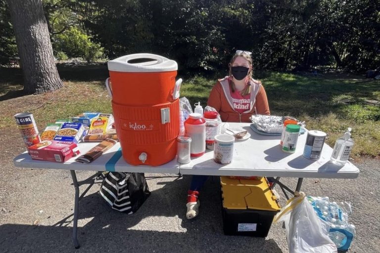 Brenna Jones sits behind a table bearing food and drinks for the unhoused and precariously housed.