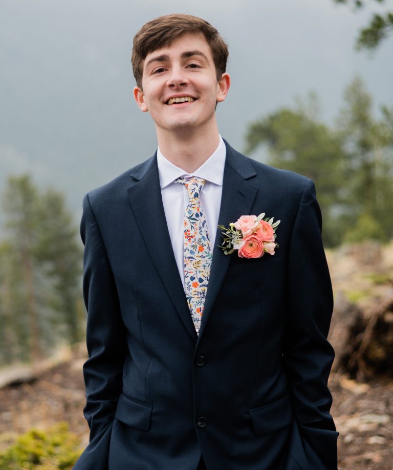 Josh Adell, wearing a dark suit and pink boutonniere, poses for a photo outside.